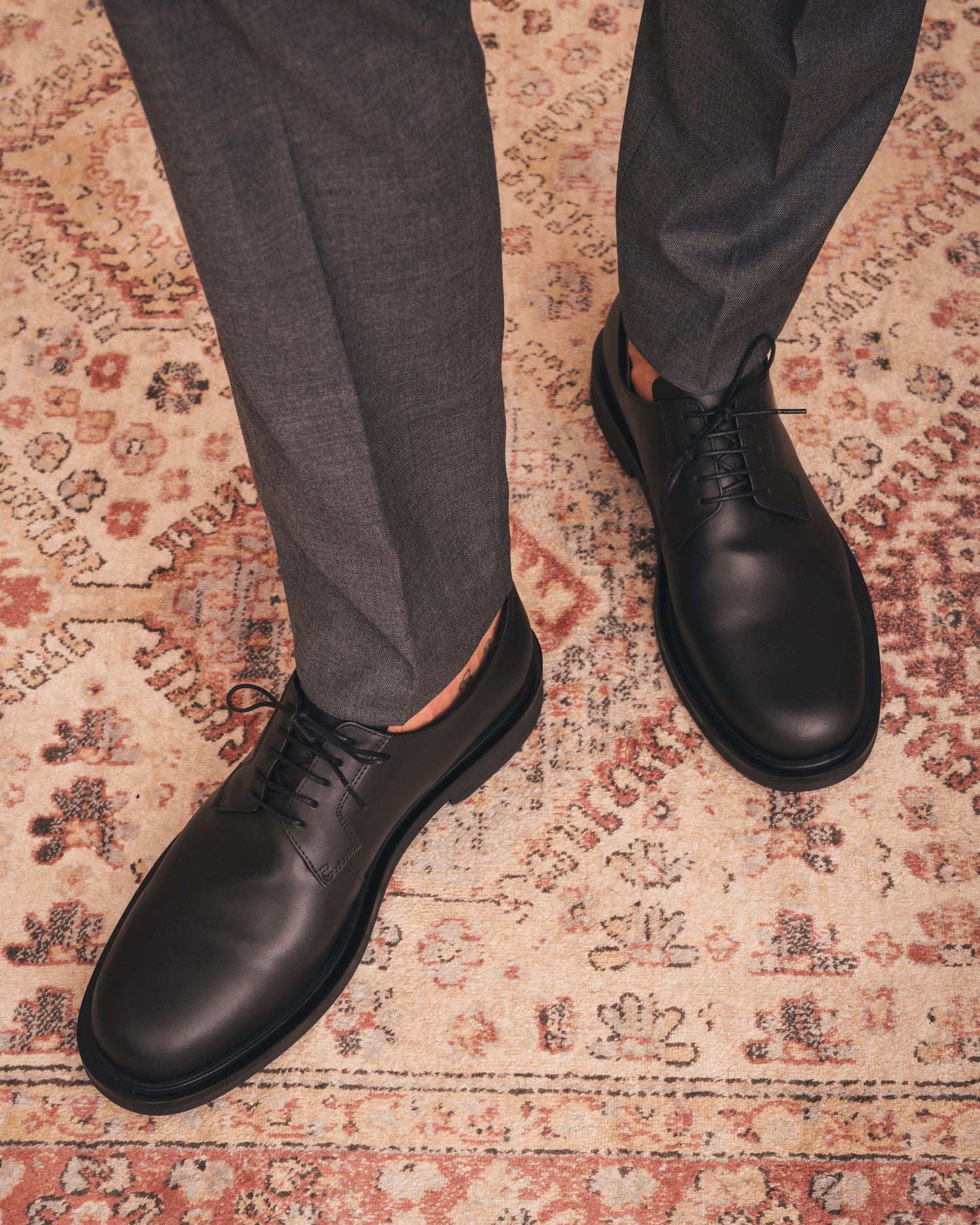 Black leather shoes worn with gray pants on a patterned rug