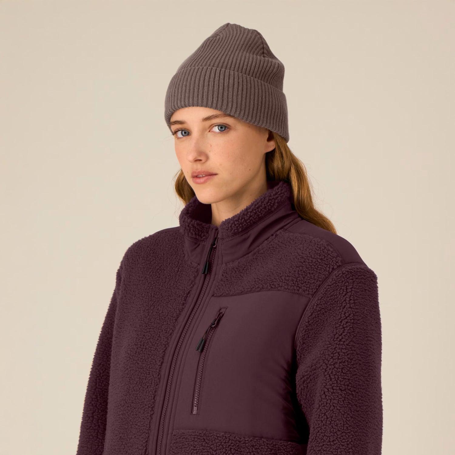 Woman wearing a vegan dark purple fleece jacket and matching beanie against a beige background