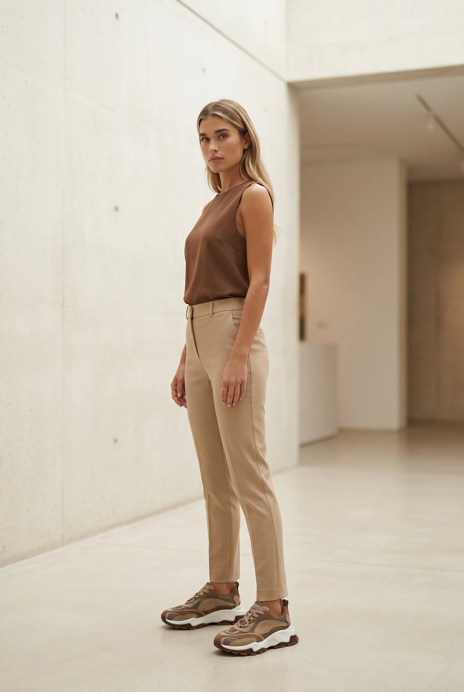 Woman wearing a brown top and beige pants standing in a minimalistic room.