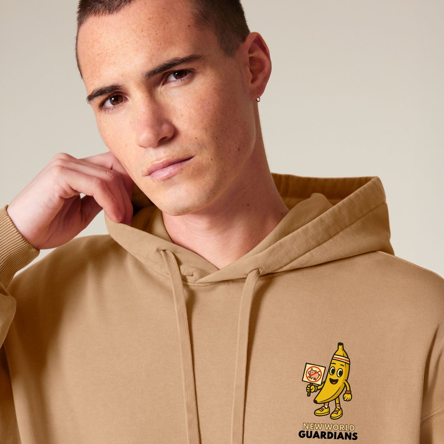 Close-up of male model wearing a latte beige NEW WORLD GUARDIANS unisex vegan hoodie with Bany the banana in a comic-style front print and the full veggie squad on the back. From the Roots for Our Future collection. Made from 100% organic cotton. Conscious vegan fashion meets sustainable streetwear style.
