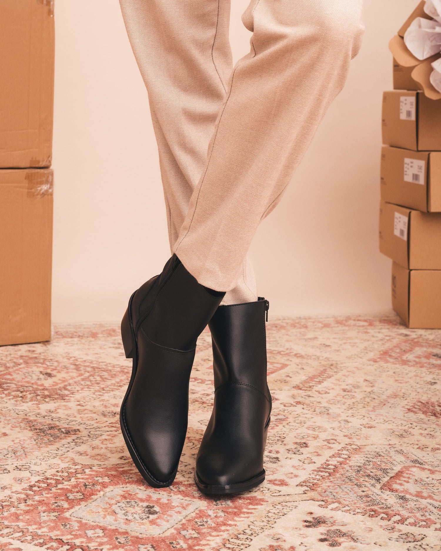 Person wearing black ankle boots with beige pants in a room with cardboard boxes.