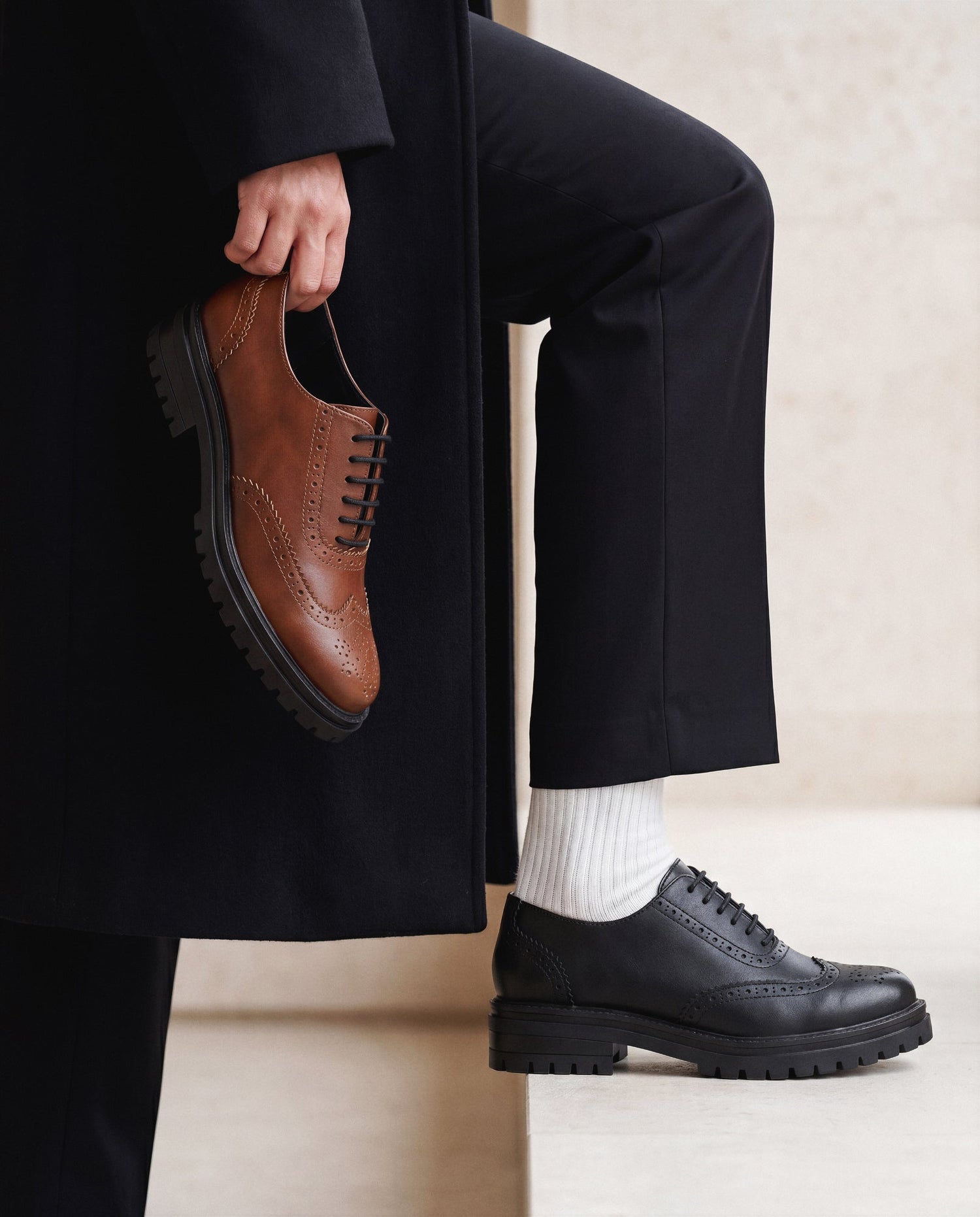 Person wearing black shoes and holding a brown shoe, with a neutral background