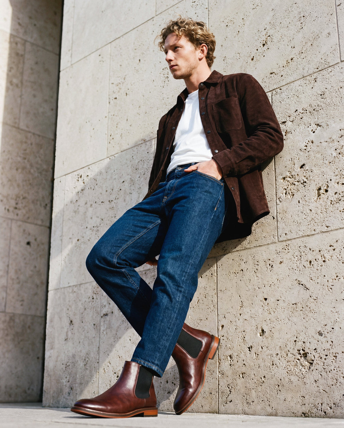 Mann in brauner Jacke, weißem Hemd, blauen Jeans und braunen veganen Stiefeln von Nae Vegan Shoes lehnt an einer Steinmauer
